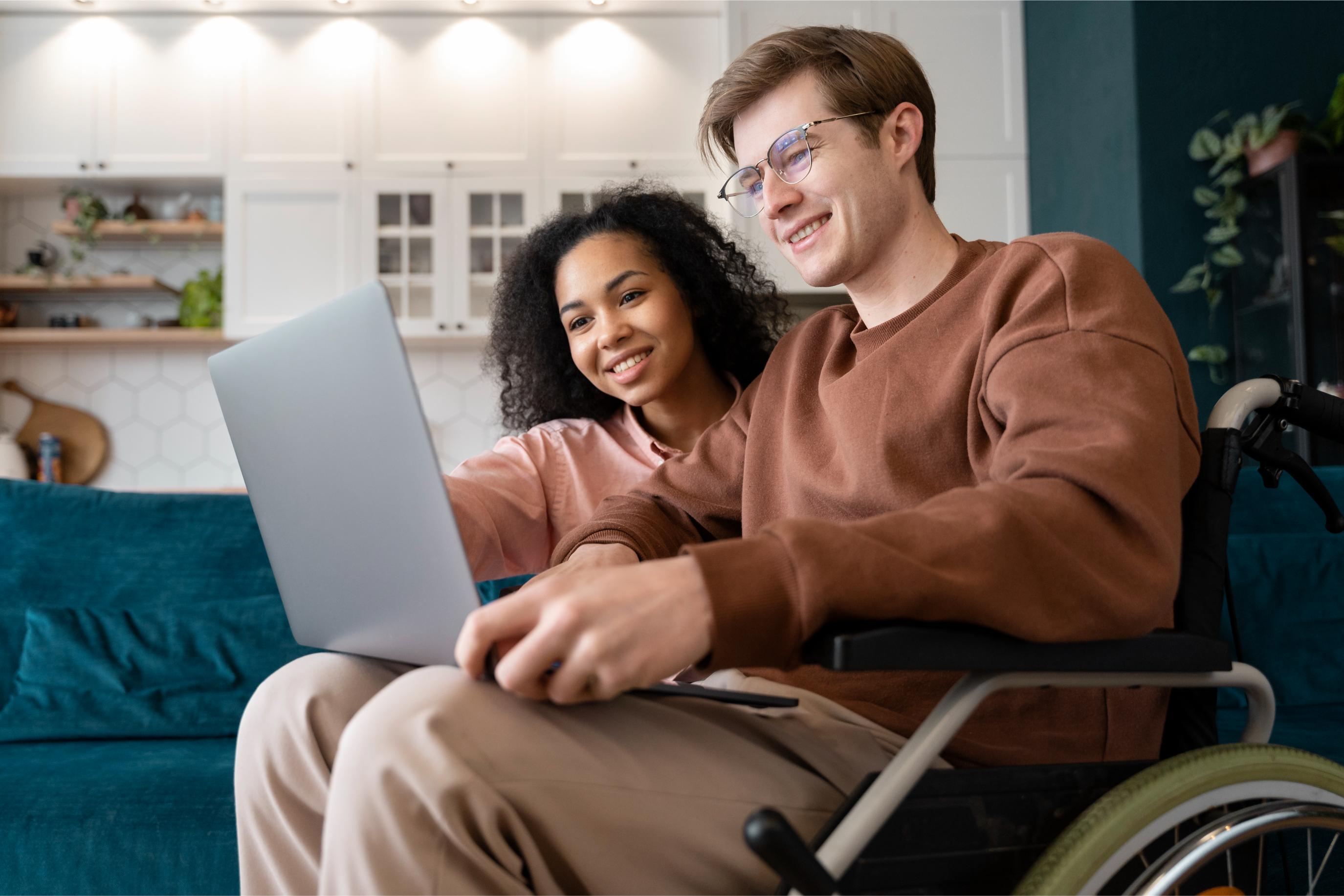A young man in a wheelchair and a woman sit closely on a couch, smiling as they look at a laptop together, illustrating a supportive and positive relationship