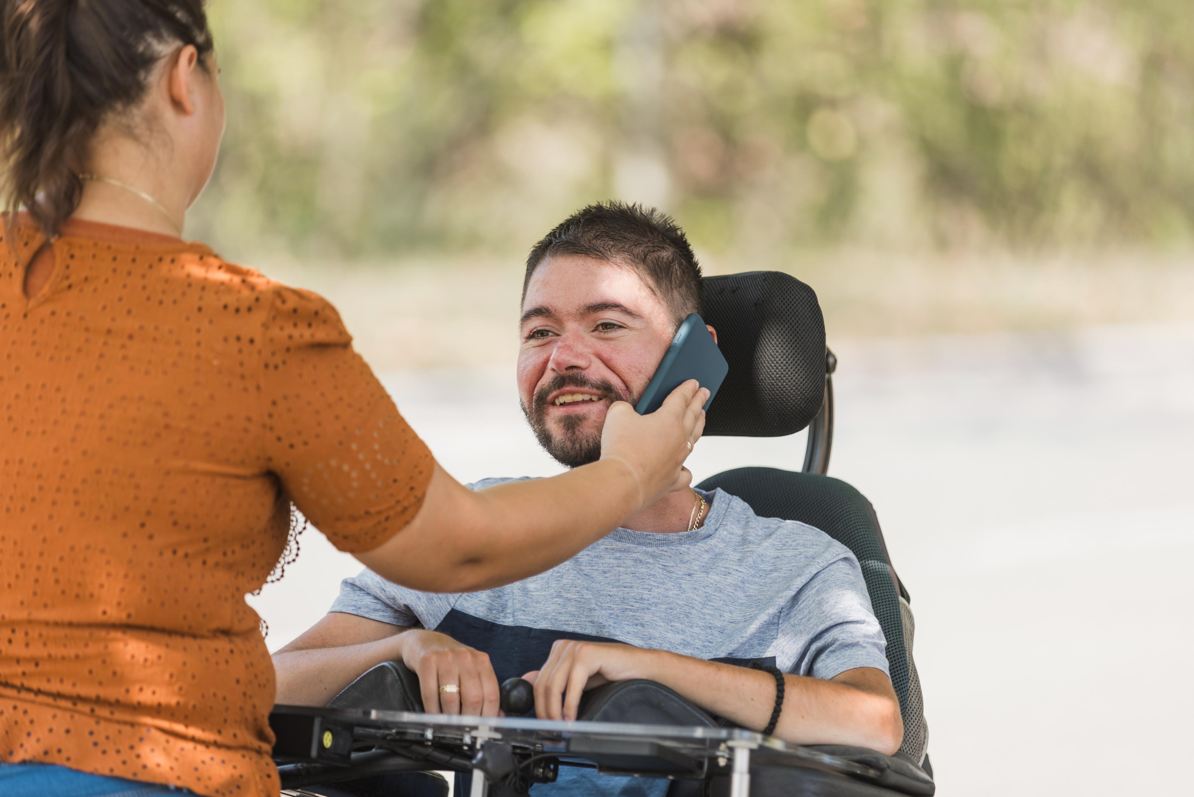 A support worker holds a smartphone to the ear of a smiling man in a wheelchair during an NDIS support visit