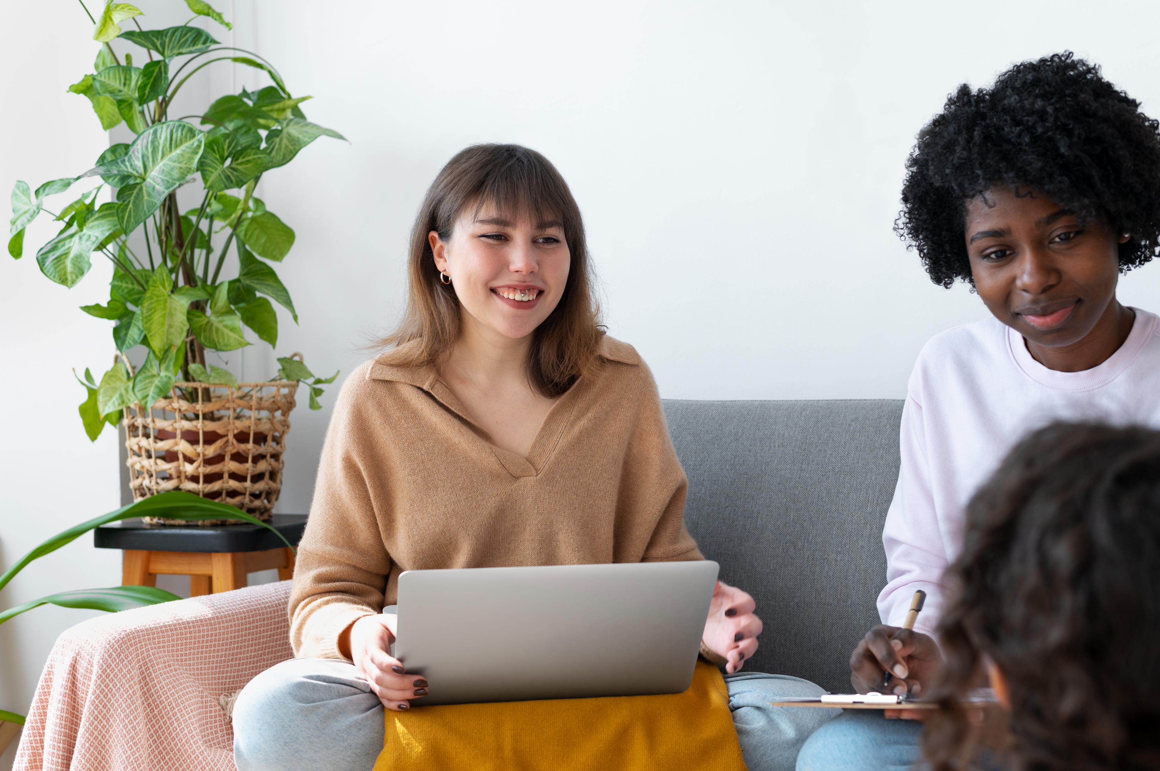 Two smiling women sit on a couch with a laptop, discussing NDIS plan variations and budget options with a consultant.