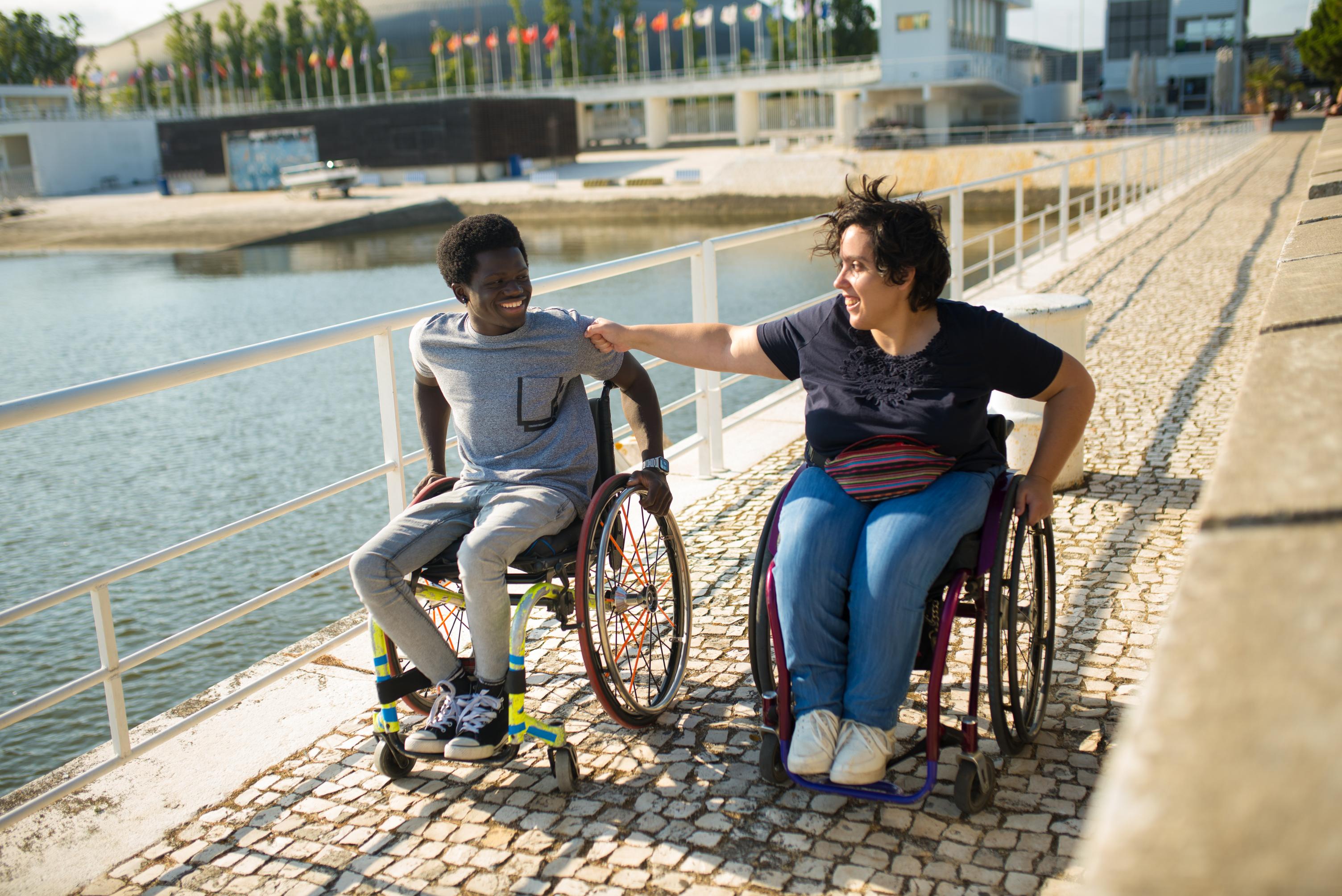 Two people in manual wheelchairs smile and talk together by a waterfront, enjoying freedom from NDIS paperwork