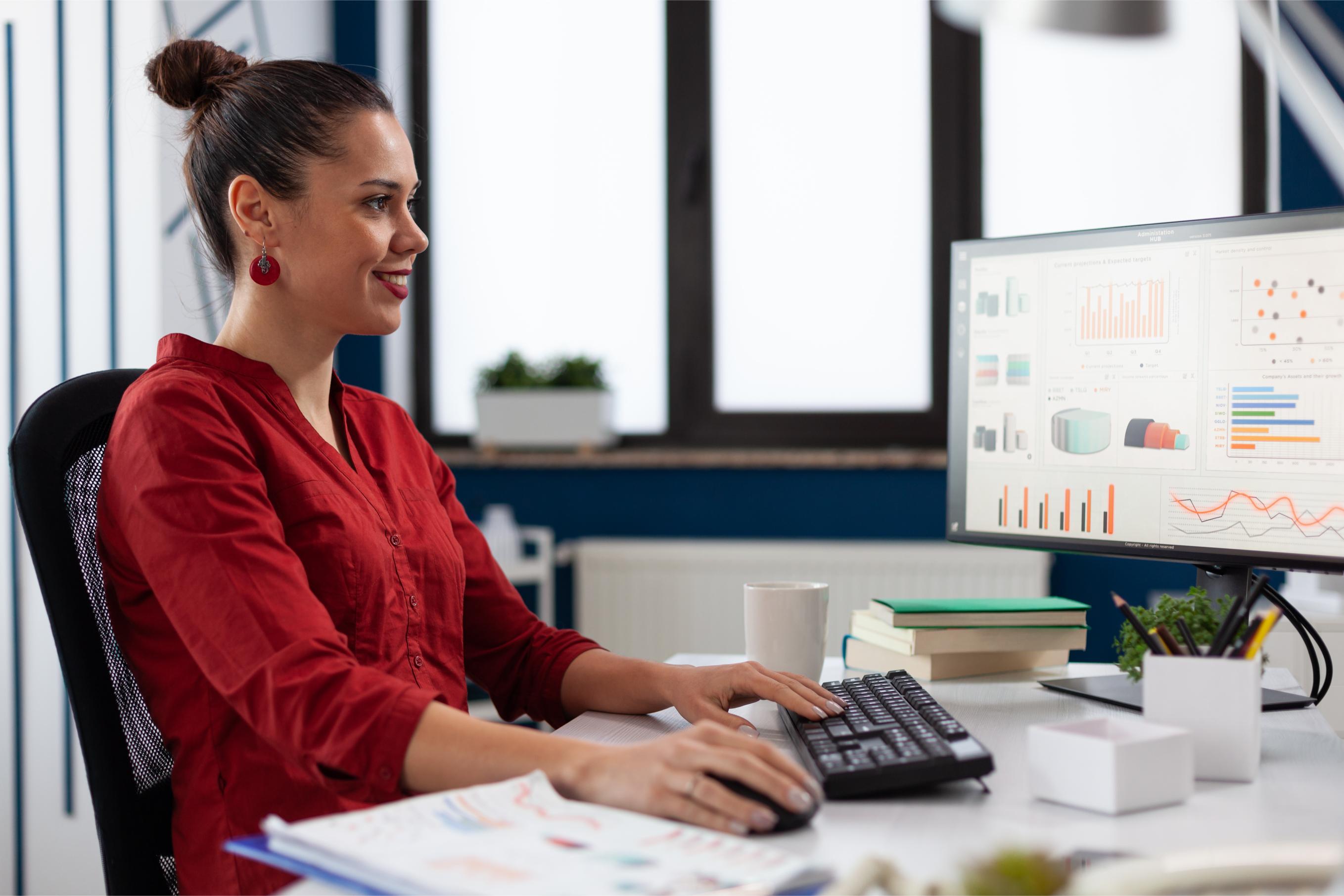 A woman smiles at an office desk while reviewing financial charts on a computer screen for NDIS plan management
