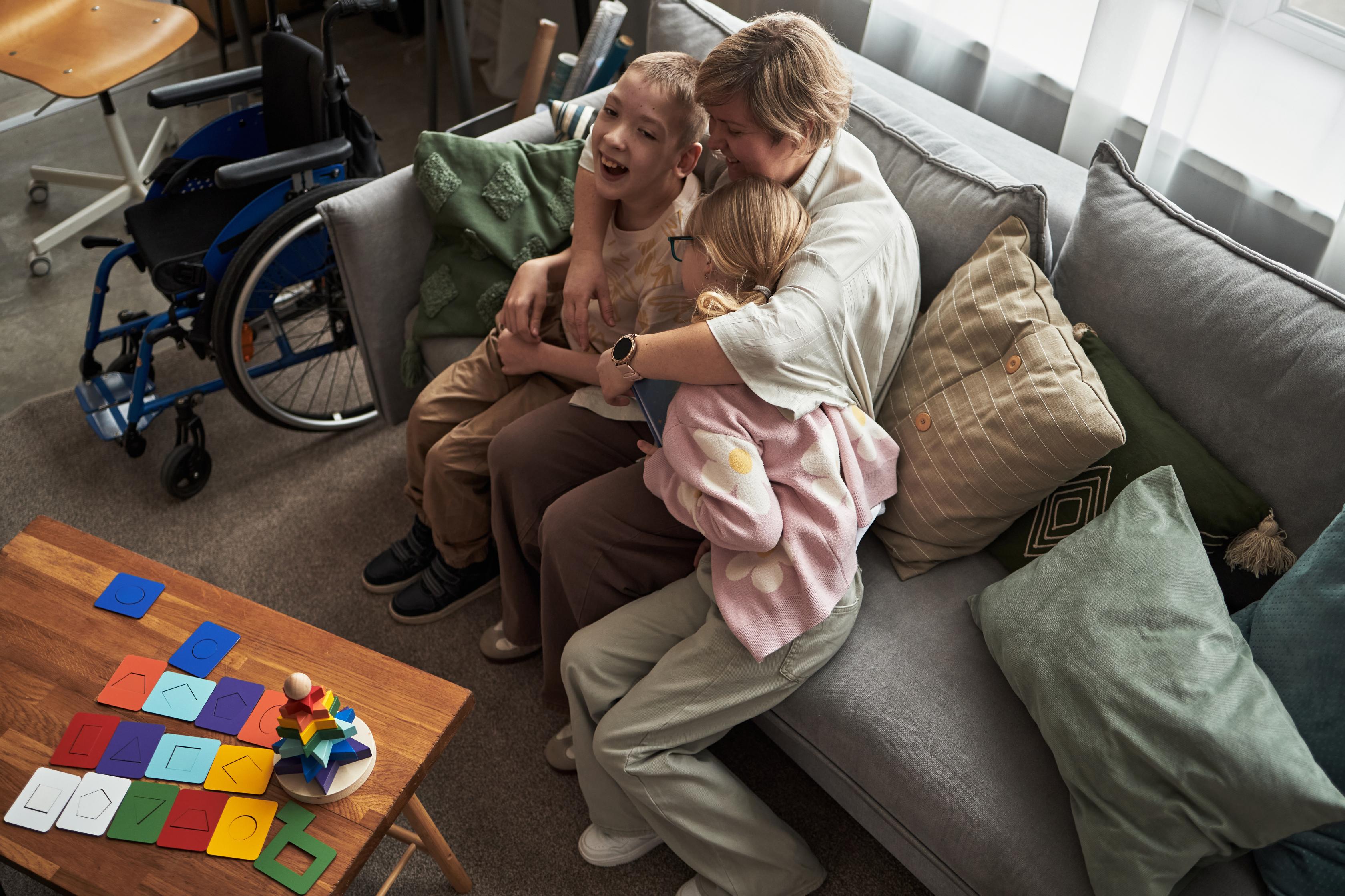 An adult provides a supportive hug to two children on a couch, with a wheelchair visible in the background, representing the importance of family relationships and in-home support