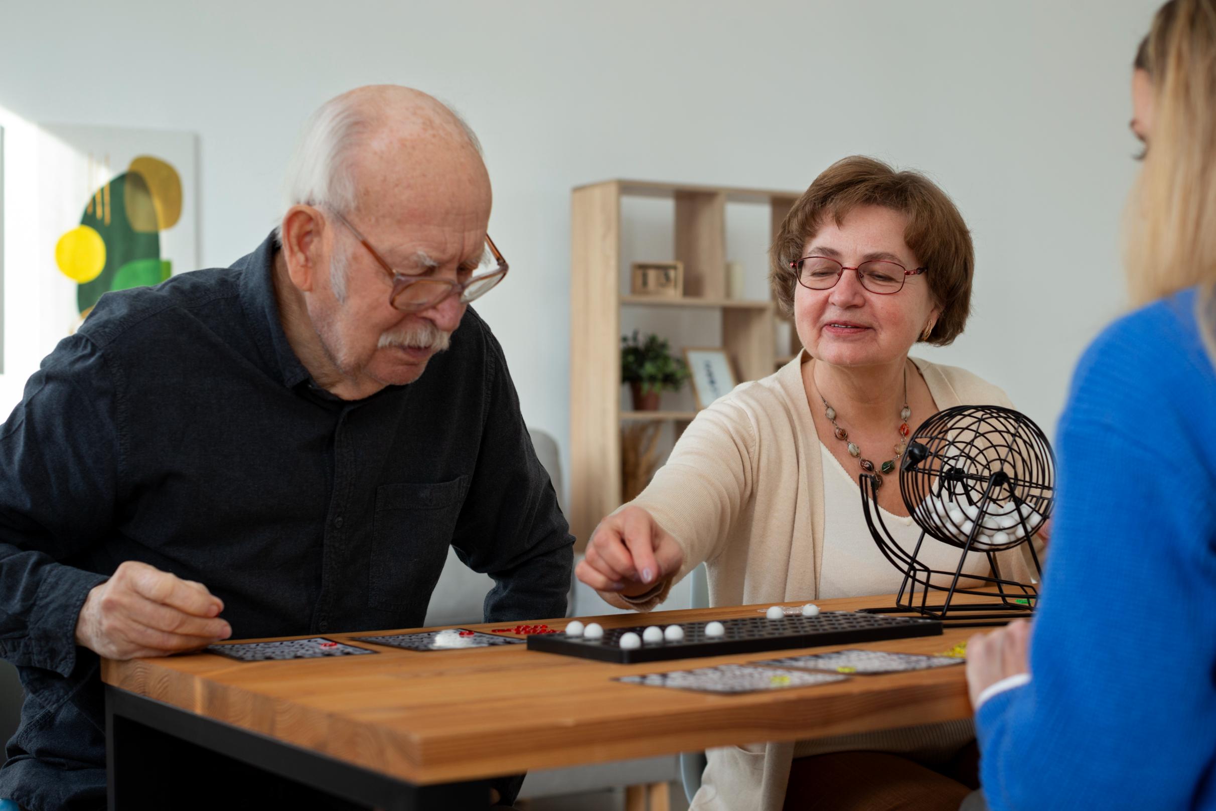 An older man and woman sit at a table playing a game of bingo with another person, illustrating participation in a social group activity
