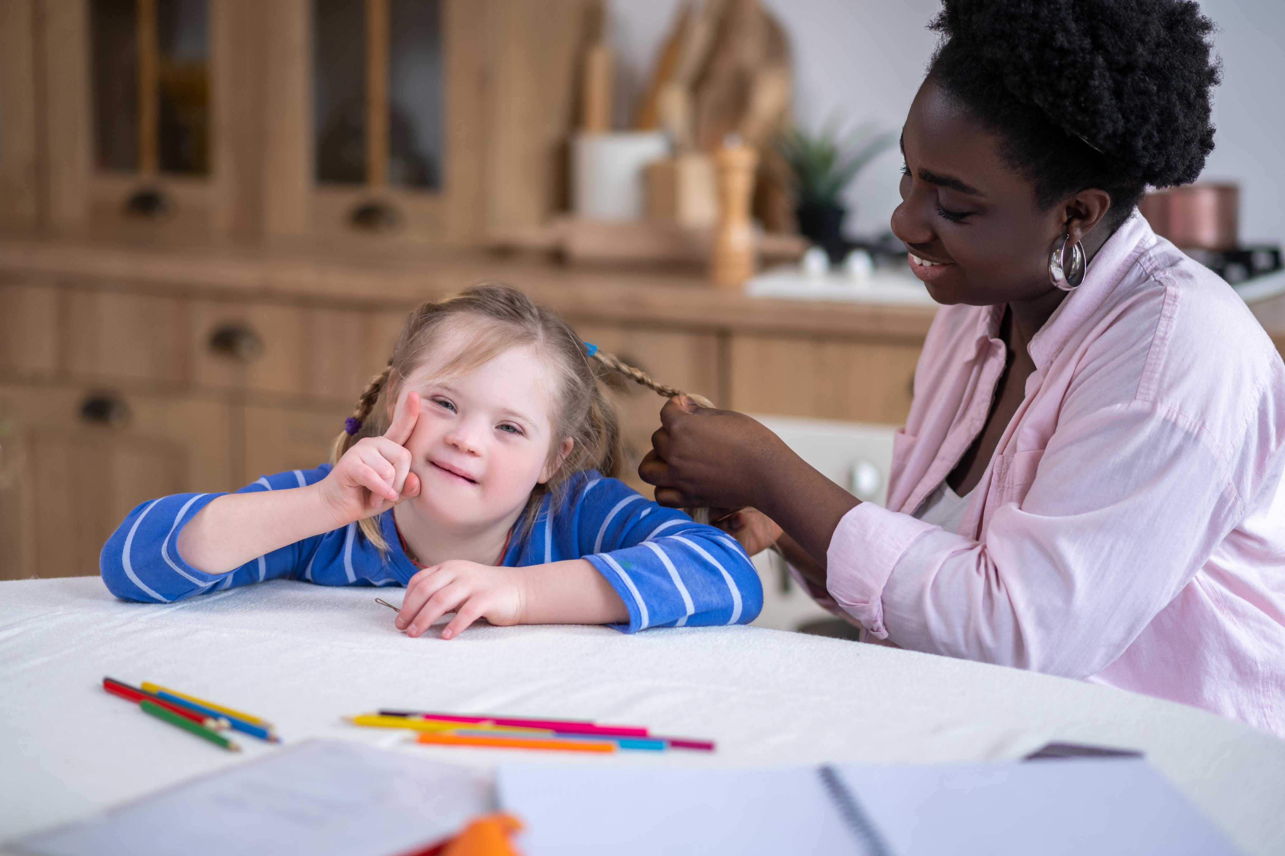 A support worker styles the hair of a smiling young girl with Down syndrome during a care service funded by the NDIS.