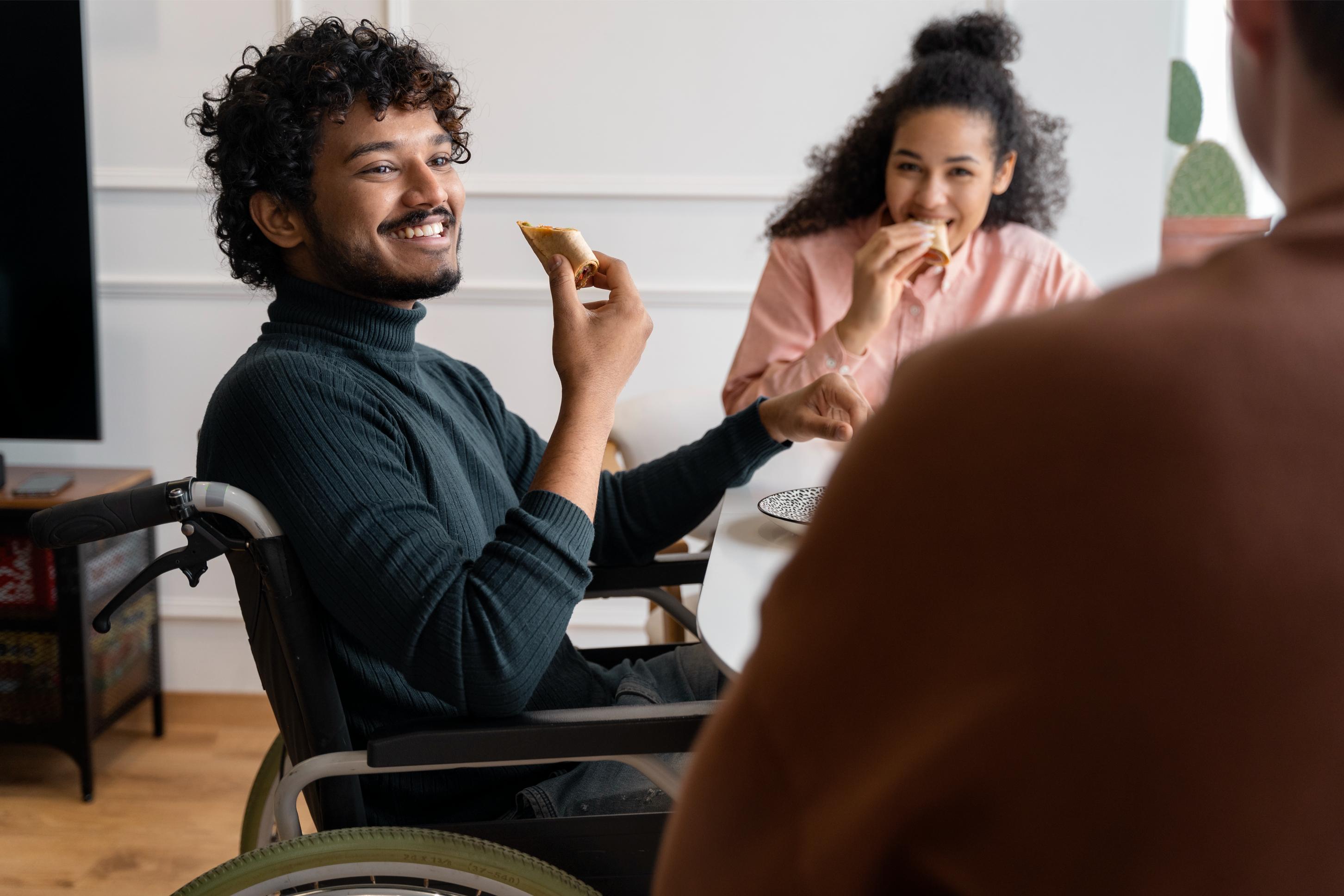 A young man in a wheelchair smiles and eats pizza at a table with two friends, demonstrating positive social connection and participation