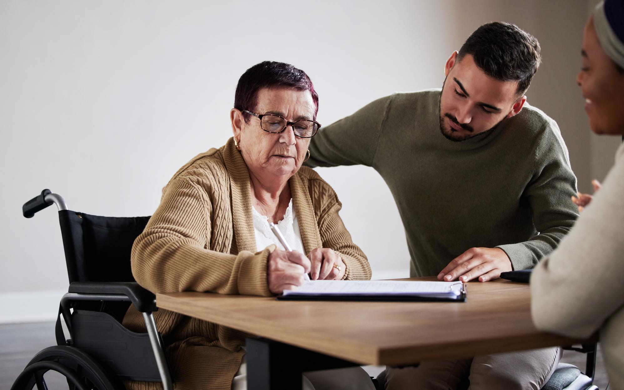 Supported by a younger man, an older woman in a wheelchair signs her new NDIS plan reassessment paperwork at a desk.