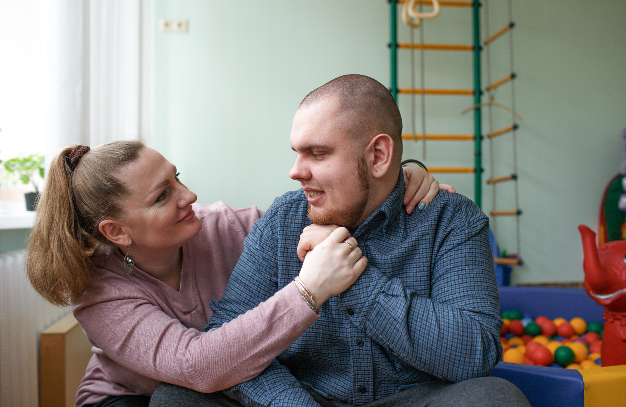A woman gently supports a young man, looking at him with care, as he engages in conversation. They are in a room with a ball pit in the background, suggesting a supportive therapy or community setting